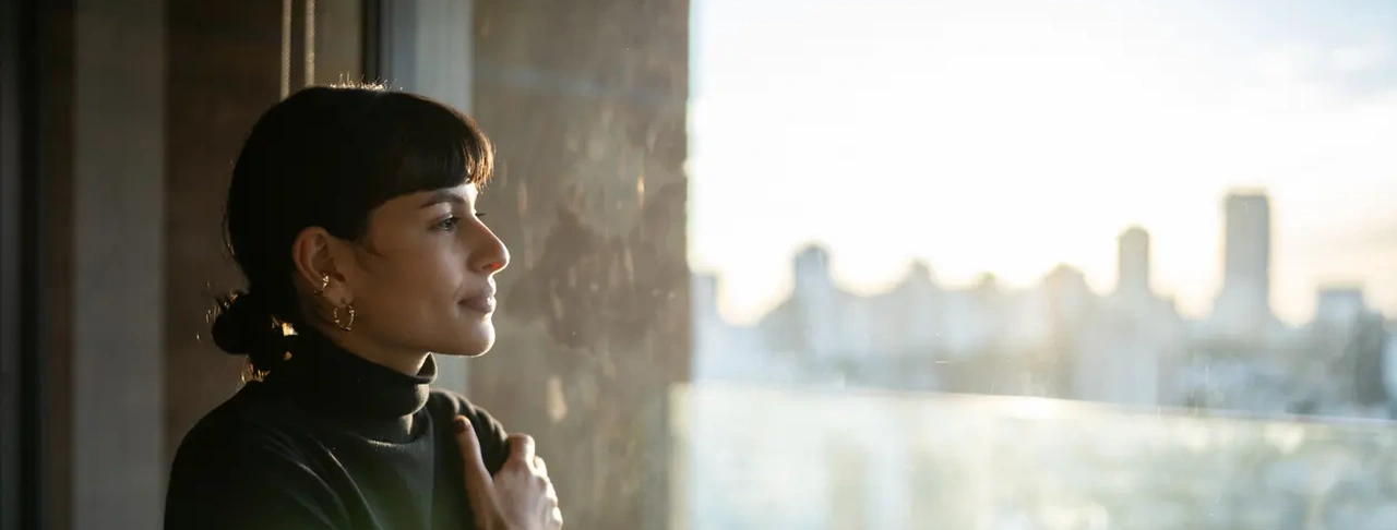 Woman standing by a sunlit window, looking outside with her hand resting on her chest.